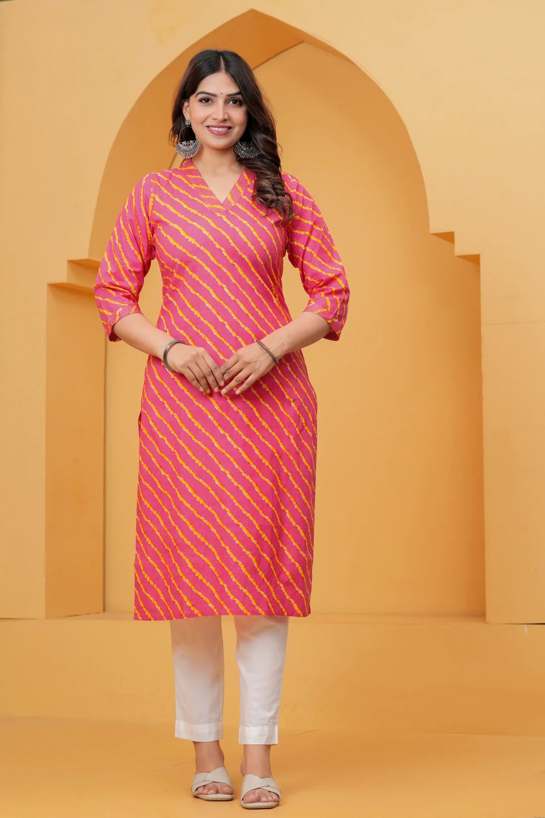 A woman posing in a pink and yellow Leheriya diagonal striped cotton kurta with white straight-cut trousers against a yellow architectural background.